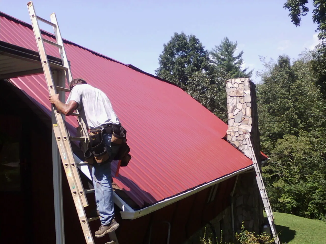 Roofer on a ladder beside a red metal roof and stone chimney — Soffit & Fascia Repair by Roofing Brothers in Marlinton